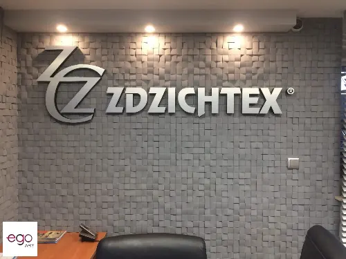 Srebrne logo przestrzenne Zdzichtex na szarej ścianie z dekoracyjną fakturą.