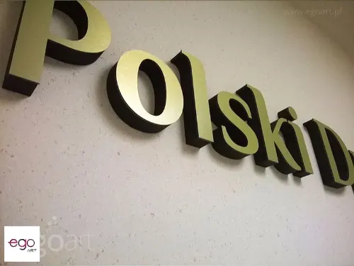 Zbliżenie złotych lic z dibondu i ciemnych boków liter zamontowanych na ścianie.