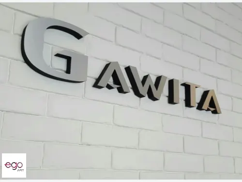 Przestrzenny napis Gawila ze srebrnym licem z dibondu na białej ceglanej ścianie.