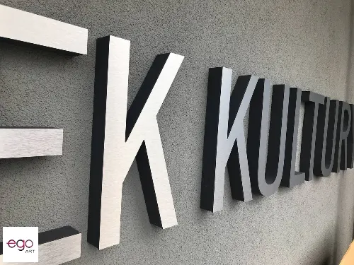 Przestrzenne litery Ośrodka Kultury ze srebrnym licem i ciemnymi bokami na elewacji budynku.