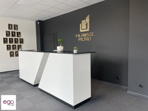 Złoty znak i napis Pierwsze Piętro nad białą ladą na tle szarej ściany.