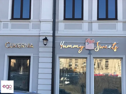 Złote napisy Cukiernia i Yummy Sweets nad witrynami lokalu na szarej elewacji.