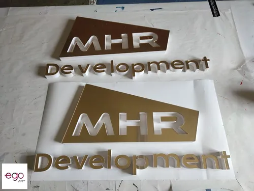 Dwa złote logotypy MHR Development przygotowane do montażu, częściowo zabezpieczone folią.