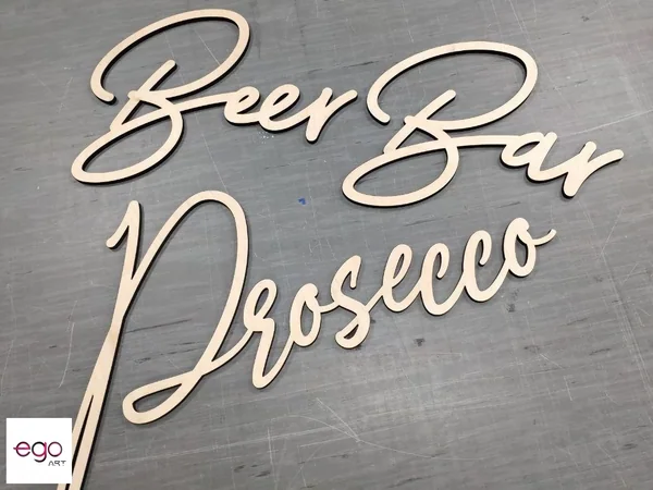 Ozdobne napisy Beer Bar i Prosecco wycięte laserowo z jasnej sklejki i ułożone na szarym tle.