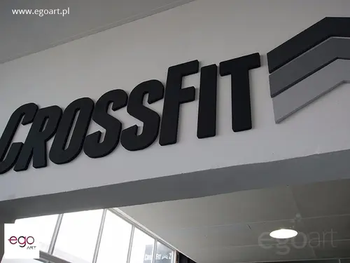 Czarny przestrzenny napis CrossFit z szarym znakiem na białej ścianie nad przejściem.