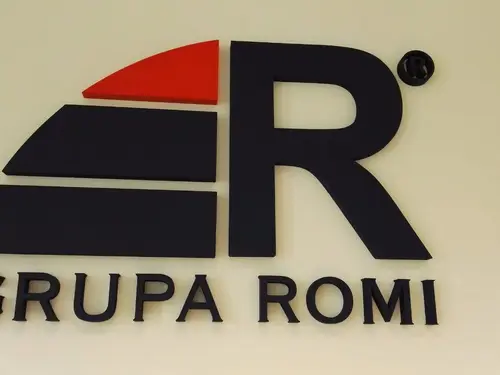 Ciemny przestrzenny znak z czerwonym akcentem i napisem Grupa Romi na jasnej ścianie.