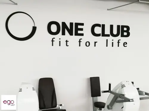 Czarny znak i napis One Club na białej ścianie nad sprzętem treningowym.