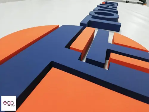 Niebieskie litery i pomarańczowe elementy przestrzennego logo pokazane przed montażem.