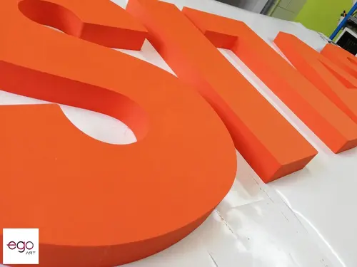 Duże pomarańczowe litery przestrzenne na stole produkcyjnym, z widoczną grubością materiału.