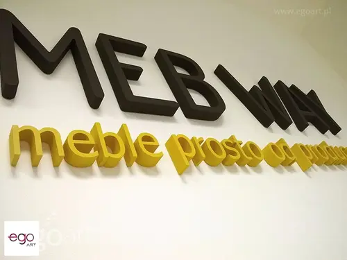 Czarne litery Mebway i żółty napis reklamowy ze styroduru na białej ścianie.