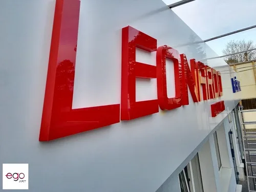 Zbliżenie czerwonych błyszczących liter przestrzennych na białym panelu reklamowym.