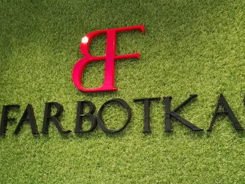 Czerwono-czarne litery Farbotka z licem z plexi na ścianie pokrytej zieloną dekoracją.