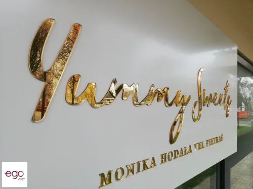 Ozdobny złoty napis Yummy z lustrzanej plexi na białym błyszczącym panelu.