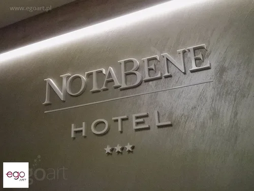 Srebrzysty napis Notabene Hotel i trzy gwiazdki na szarej ścianie wewnątrz budynku.