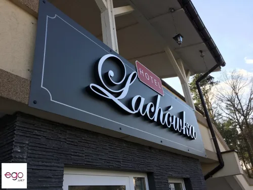 Biały ozdobny napis Hotel Radawka z ciemnymi bokami na szarym szyldzie nad wejściem.