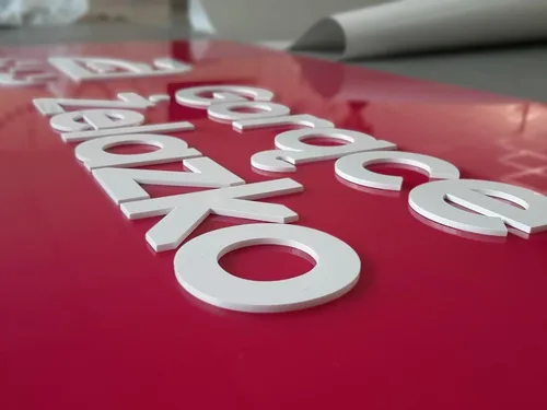 Białe litery z plexi i elementy znaku reklamowego rozłożone na czerwonym panelu.