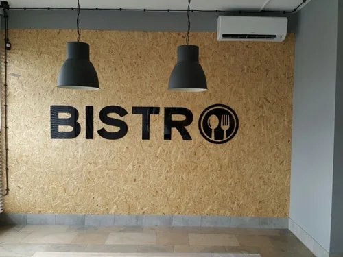 Czarne litery Bistro i okrągły piktogram sztućców z plexi na ścianie z płyty OSB.