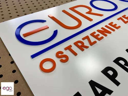 Niebieskie, pomarańczowe i czarne litery z plexi na białej tablicy reklamowej.