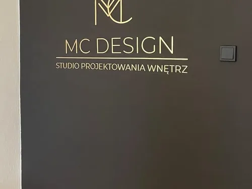 Złoty znak i napis MC Design z lustrzanej plexi na ciemnej ścianie.