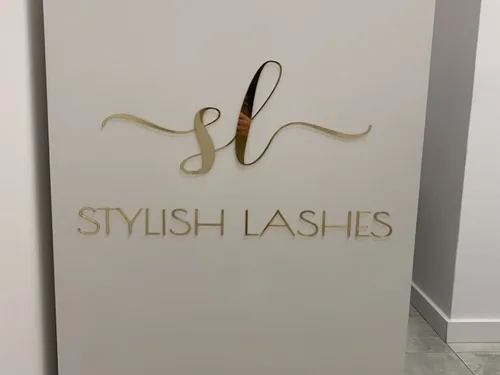 Delikatny złoty znak i napis Stylish Lashes z lustrzanej plexi na jasnej ścianie.