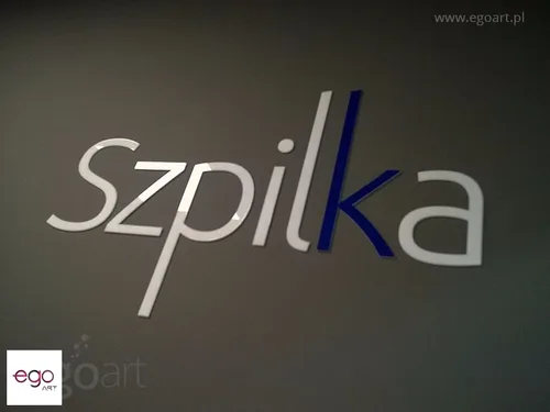 Jasnoszary napis Szpilka z granatowym akcentem na szarej ścianie.