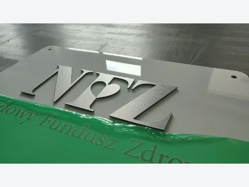 Srebrne litery NFZ ze szczotkowanego dibondu na zielono-białej tablicy.