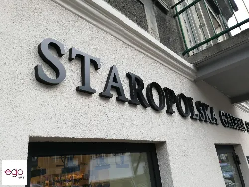Zbliżenie czarnych liter przestrzennych na jasnej elewacji restauracji.