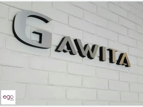 Przestrzenny napis Gawila ze srebrnym licem z dibondu na białej ceglanej ścianie.