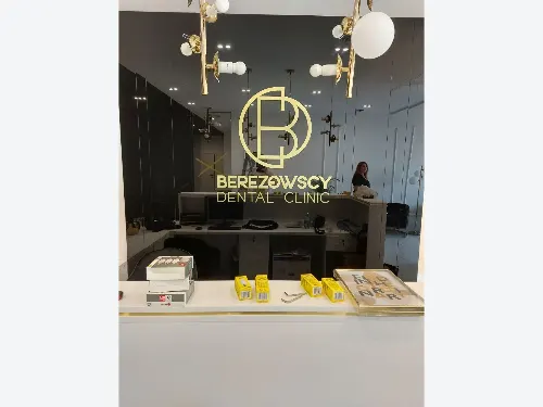 Złote logo Berezowscy Dental Clinic na ciemnym tle za białą ladą recepcyjną.