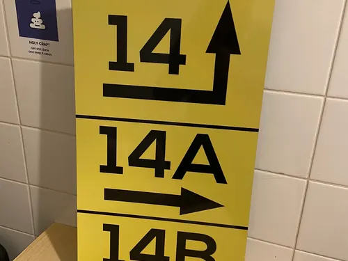 Żółta tablica z numerami 14, 14A i 14B oraz czarnymi strzałkami, wykonana z użyciem folii odblaskowej, w zwykłym oświetleniu pomieszczenia.