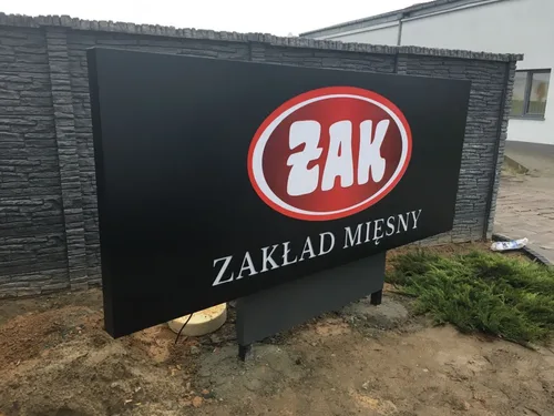 Czarna tablica z czerwono-białym logo Żak i napisem Zakład Mięsny na dwóch słupkach.