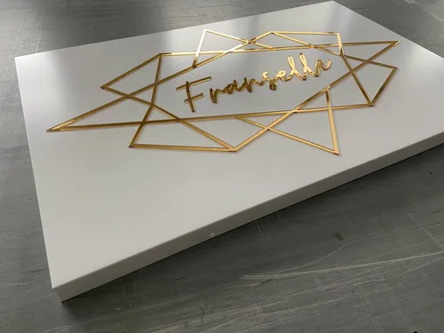 Biały panel reklamowy z wypukłym złotym napisem i geometryczną ramką wokół logo.