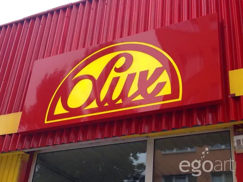 Czerwony kaseton reklamowy z żółtym logo Lux na elewacji sklepu Społem.