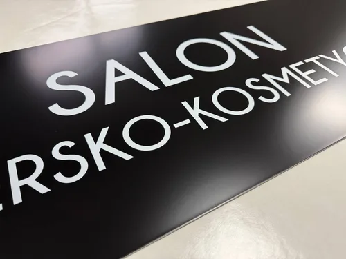 Czarny szyld salonu fryzjersko-kosmetycznego z białym napisem wykonanym nadrukiem UV.