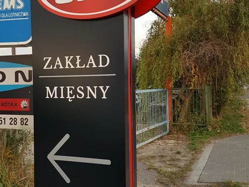 Pionowy czarny szyld ze strzałką i wypukłym napisem Zakład Mięsny, zamontowany przy wjeździe.