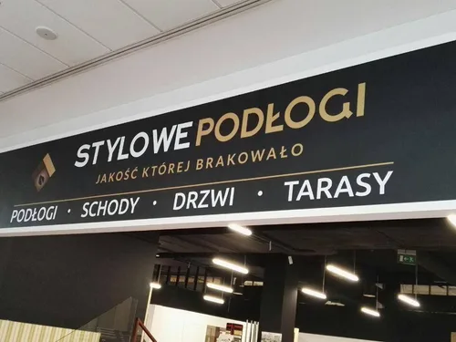 Długi czarny szyld z PCV z nadrukiem nazwy sklepu i oferty podłóg, schodów, drzwi oraz tarasów.