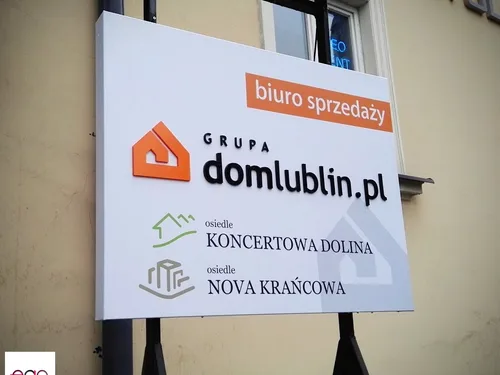 Biały szyld na zaginanym dibondzie z wypukłym pomarańczowym znakiem i czarnym logo biura sprzedaży.