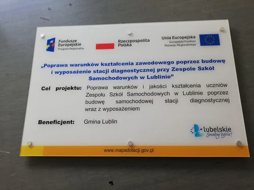 Tablica z plexi z logotypami Funduszy Europejskich i informacją o dofinansowaniu projektu, zamontowana na dystansach.