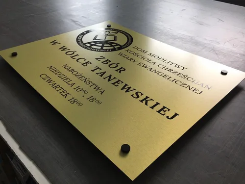 Zbliżenie złotej tablicy informacyjnej z czarnym nadrukiem i czarnymi mocowaniami dystansowymi.