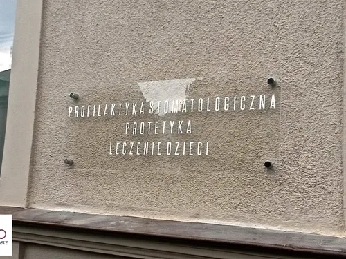 Przezroczysta tabliczka z białymi napisami o usługach stomatologicznych na srebrnych dystansach.