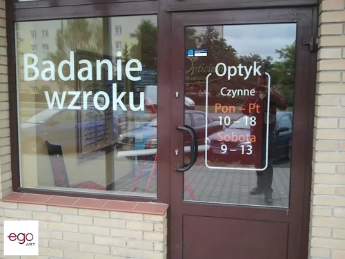 Białe litery i godziny otwarcia na szybie salonu optycznego